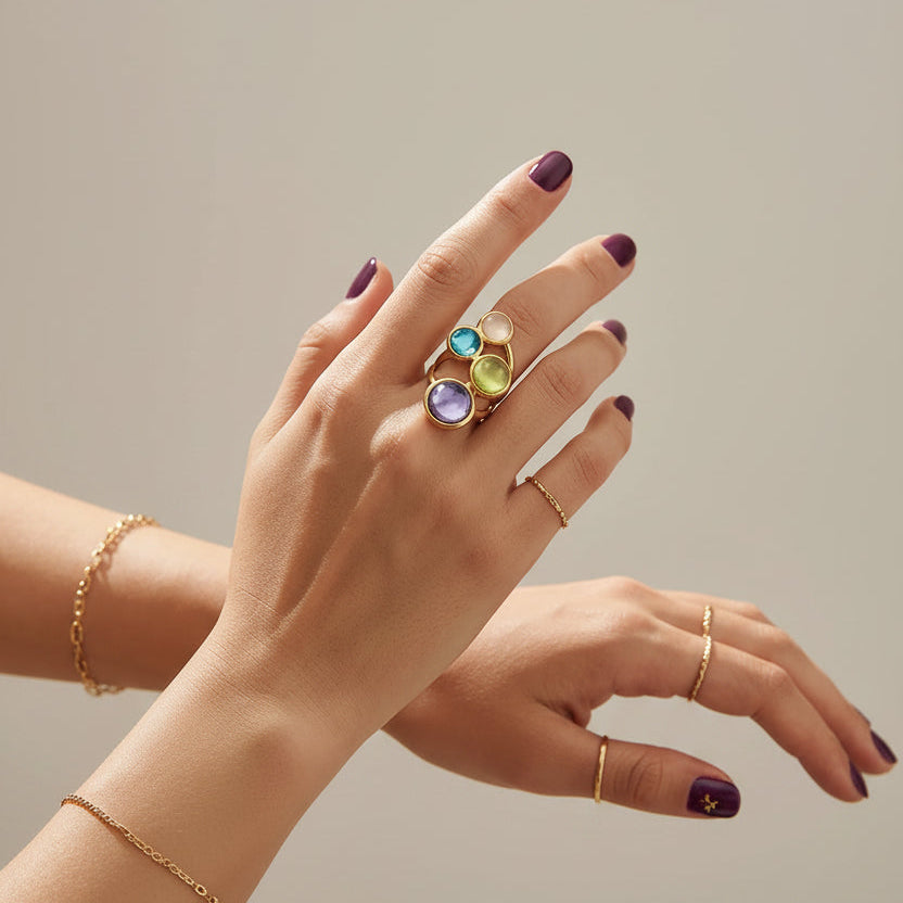 A close-up lifestyle shot of a hand wearing a gold ring set with colorful zirconia stones (blue, green, and purple) in a warm, styled indoor setting with plants and a glass of lemon tea. A brand logo overlay for 'E.