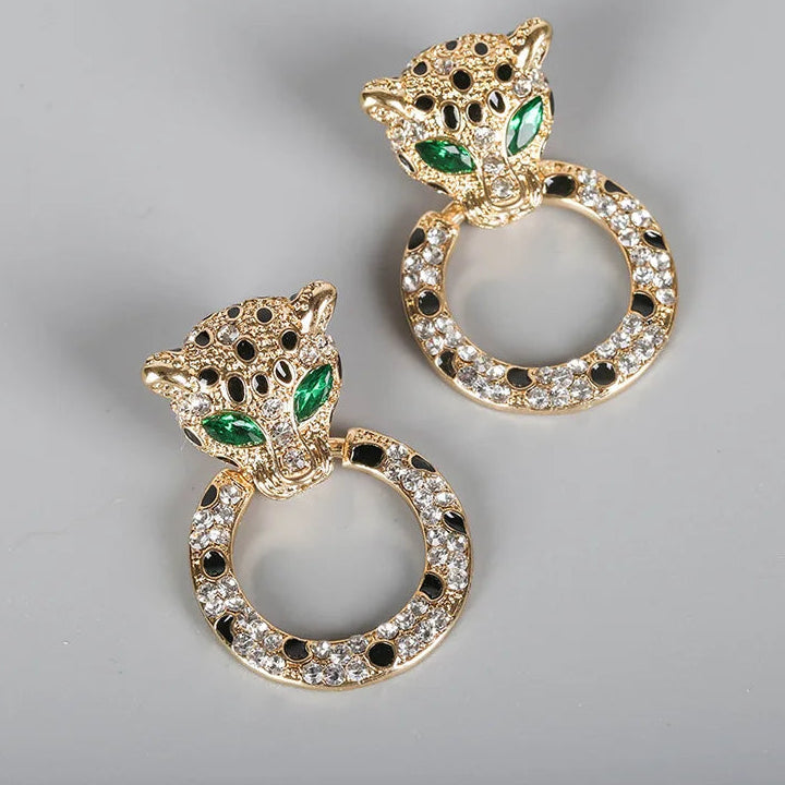 A pair of gold-toned leopard drop earrings with circular ring charms, embellished with clear rhinestones, black stones, and green emerald-cut eyes. The brand name 'lennik' is visible in the top left corner.