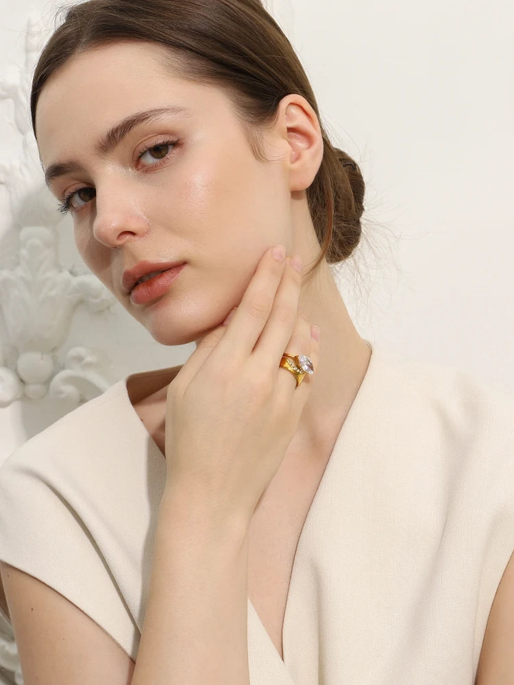 A close-up lifestyle shot of a woman with her hair in a low bun, wearing a gold ring with a central clear gemstone on her finger, set against a soft, light-colored background.
