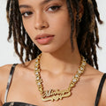 A close-up of a model wearing a gold nameplate necklace with heart details and clear rhinestones. The nameplate is personalized with the name 'Michaya' in cursive script, set against a clean white background.