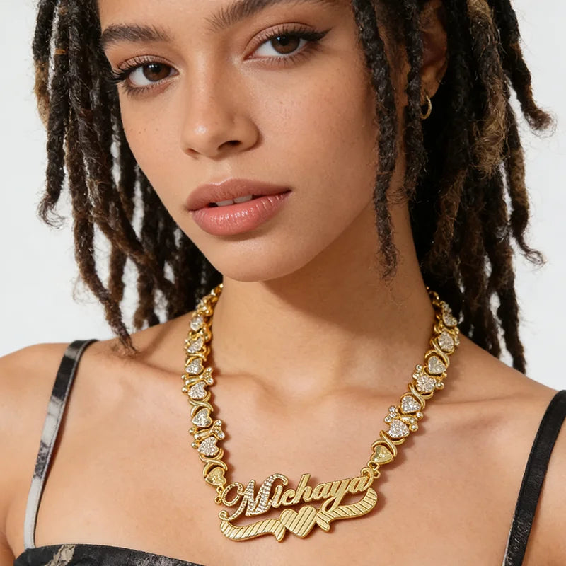 A close-up of a model wearing a gold nameplate necklace with heart details and clear rhinestones. The nameplate is personalized with the name 'Michaya' in cursive script, set against a clean white background.