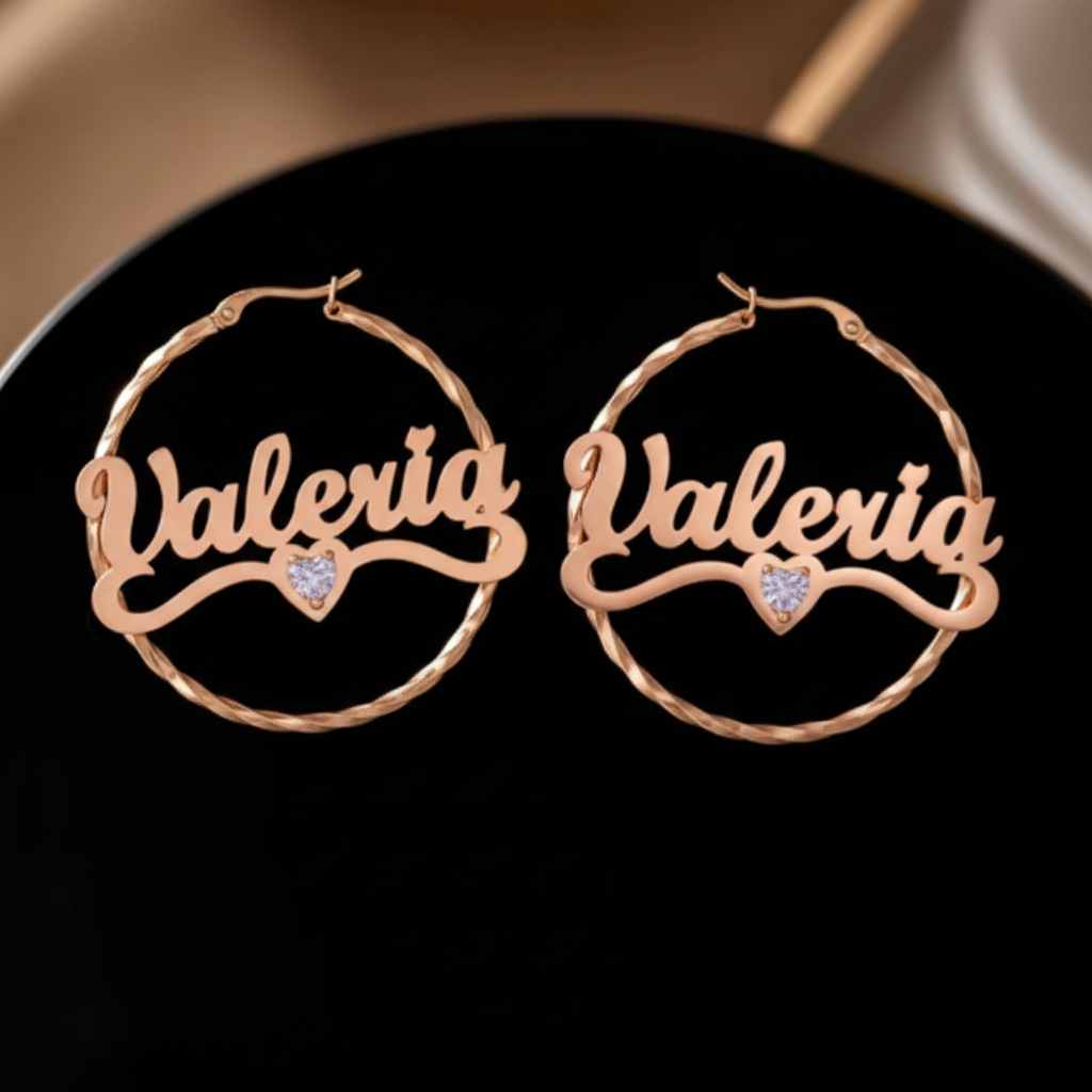 A pair of gold hoop earrings with the name 'Valeria' written across them in cursive, each featuring a small heart-shaped crystal at the center, displayed on a black surface.